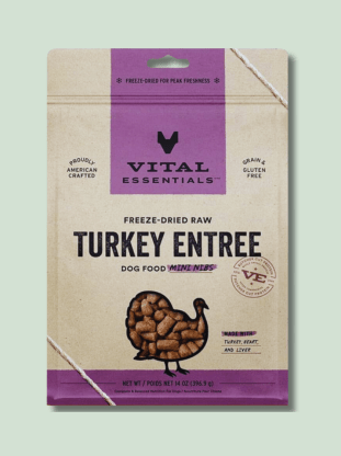 A package of Vital Essentials Freeze Dried Food raw turkey entree dog food mini nibs, marked as grain and gluten free, crafted in America, weighing 14 ounces. The package is predominantly purple and sand-colored with an illustration of the product at the bottom.