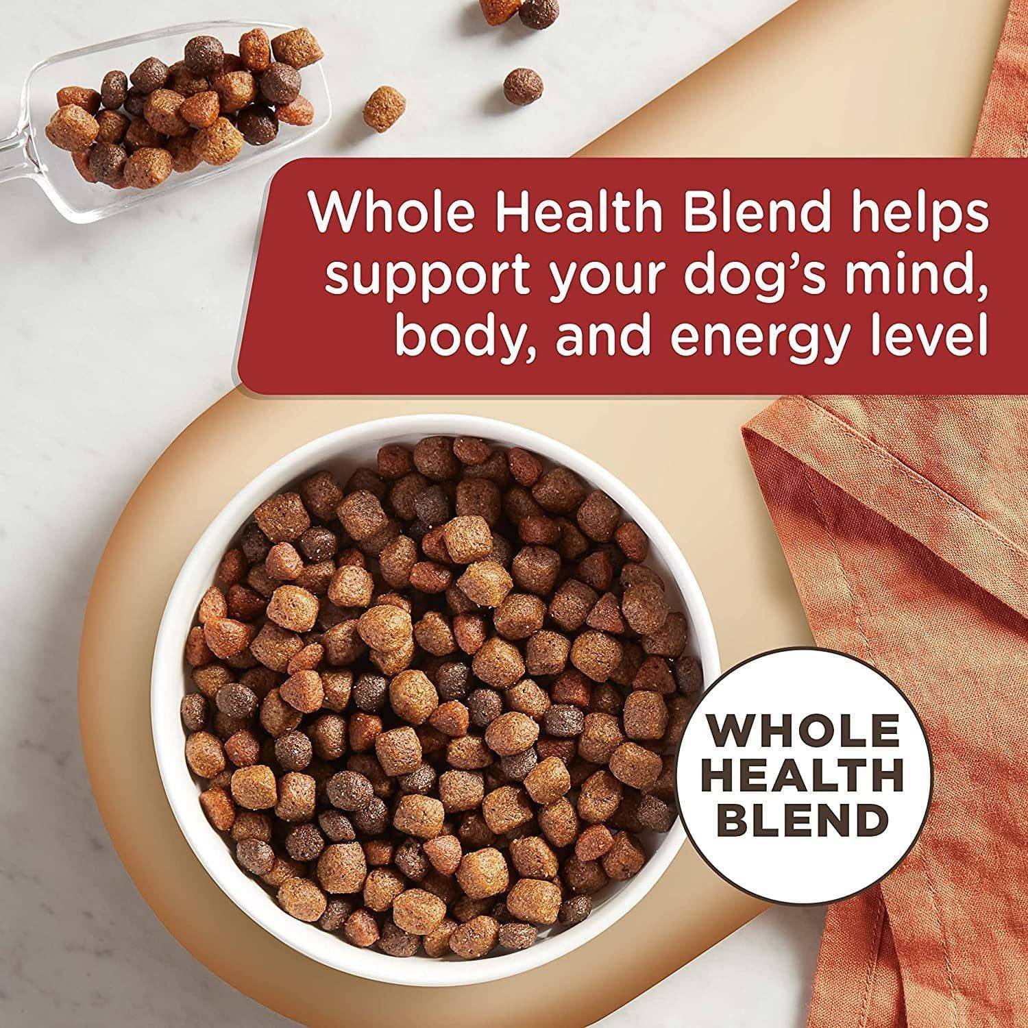 A white bowl filled with brown, round dog food kibbles on a white and beige background with a maroon and white label that reads "Whole Health Blend helps support your dog's mind, body, and energy level" and a smaller white circle label that says "WHOLE HEALTH BLEND". A clear scooper with more kibbles is also visible in the background.