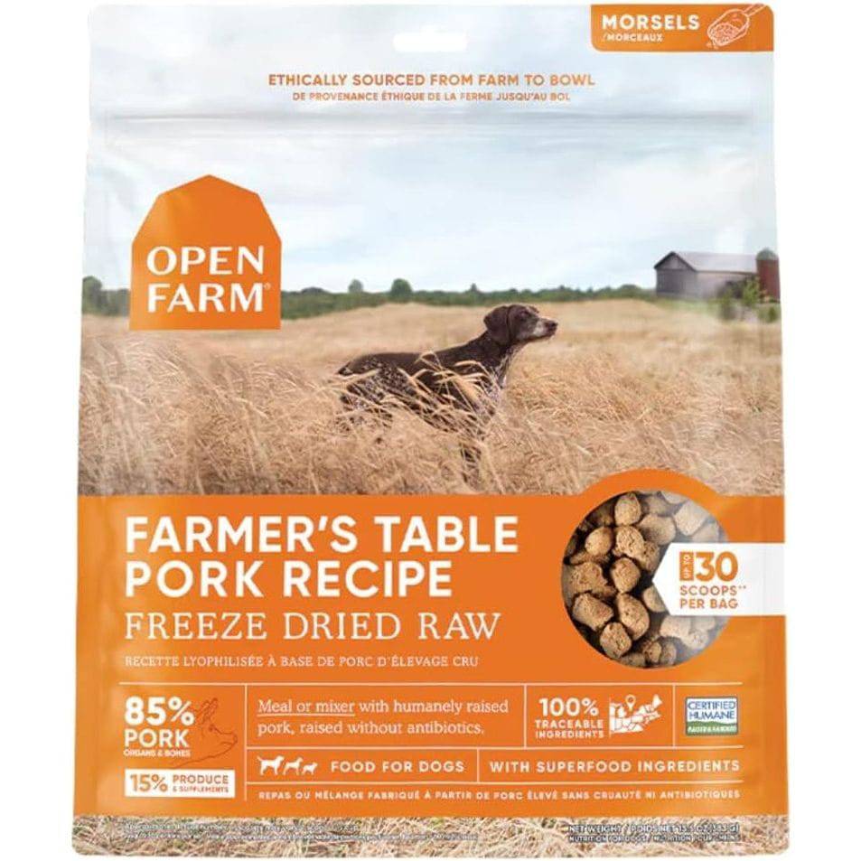 A package of Open Farm Farmer's Table Pork Recipe freeze-dried raw dog food, featuring a photo of a black dog lying in a wheat field under a cloudy sky, and information highlighted about the product containing 85% pork, humanely raised without antibiotics, and 30 scoops per bag.