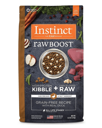 A package of Instinct Raw Boost high-protein dog food featuring freeze-dried raw kibble. The front of the bag displays a bowl of kibble surrounded by raw duck, sliced apples, carrots, and herbs. Text emphasizes that the primary ingredient is cage-free duck and the recipe is grain-free. The packaging is labeled suitable for all life stages and it is made in the USA.