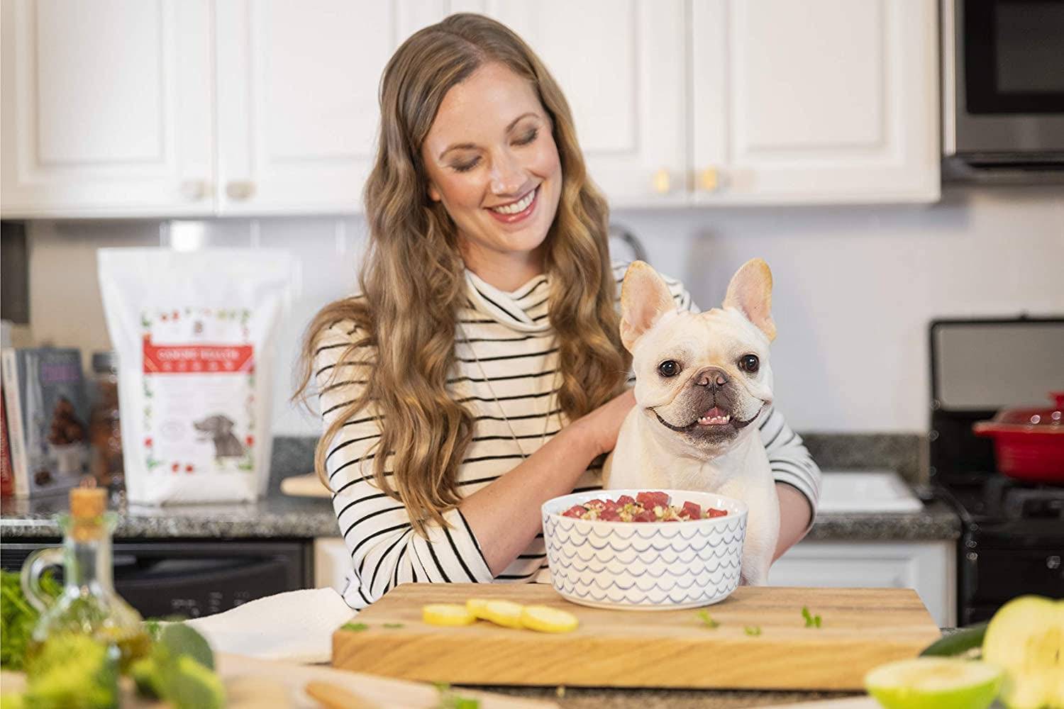 A woman holding a dog in a kitchen, both looking at a bowl of food on the counter.