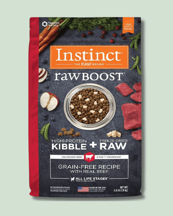 A package of "Instinct Raw Boost" high-protein kibble and freeze-dried raw beef dog food showing contents clearly displayed alongside fresh vegetables, garlic cloves, and berries on a grey surface. The bag is predominantly red and features the product logo along with details about the grain-free recipe for all life stages.